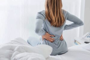 Woman sitting on edge of bed holding her arms to her lower back